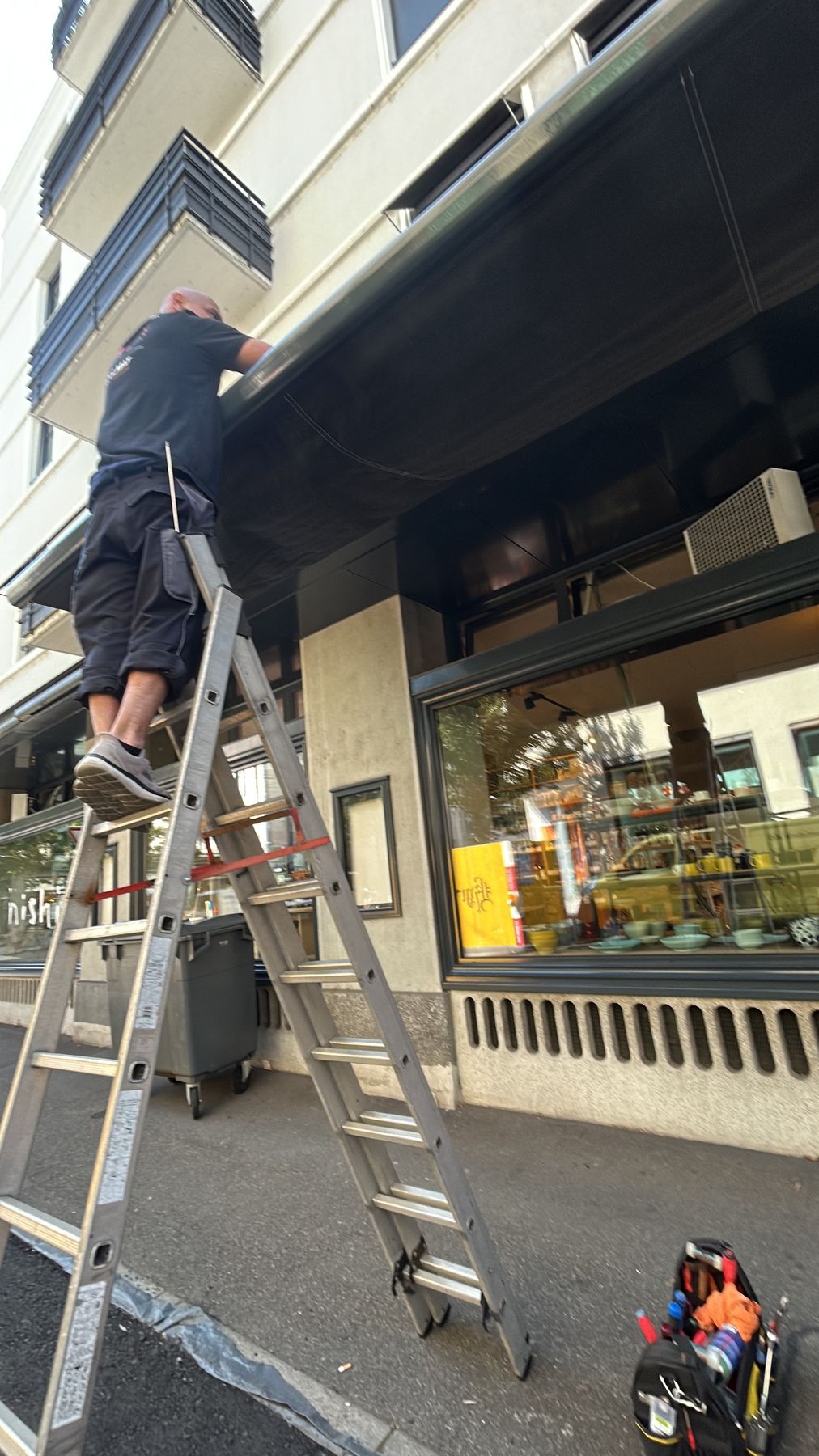 Monteur auf einer Leiter bei der Reparatur einer Markise vor einem Geschäft